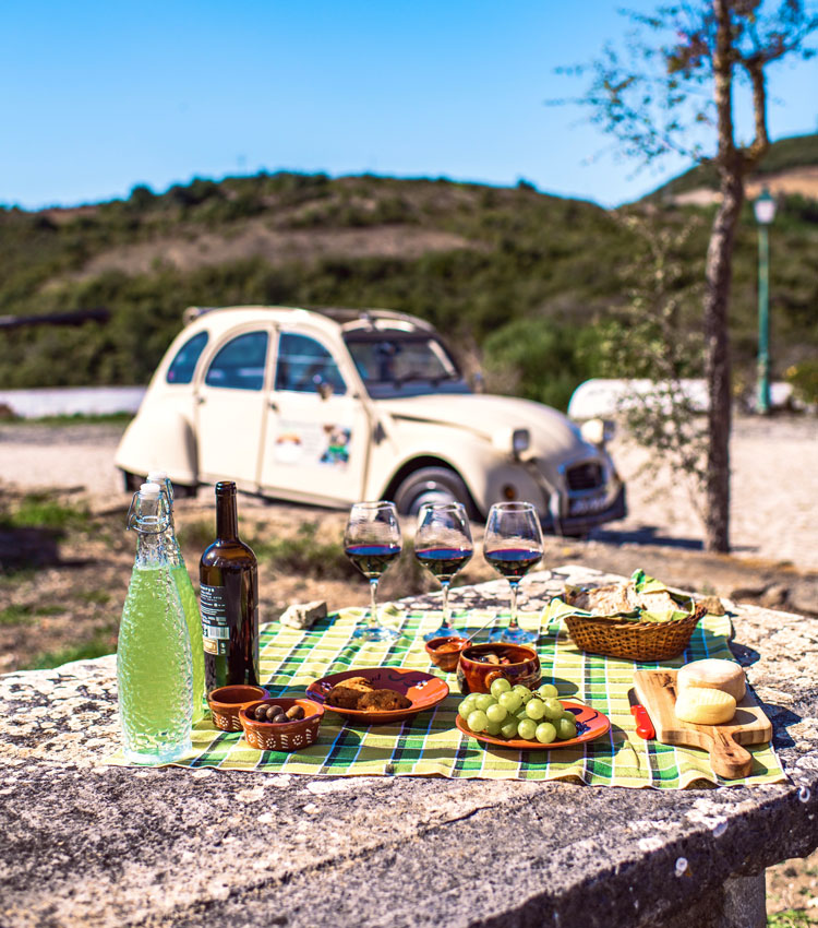 A vintage car parked beside a picnic of local wine, grapes and cheese on a stone table in the Colares countryside