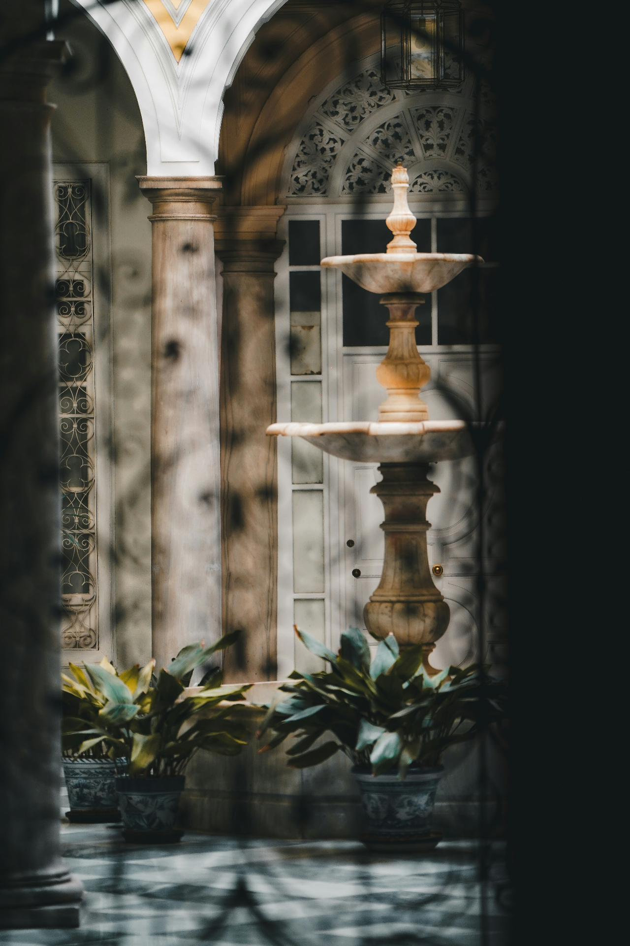 A quiet courtyard glimpsed through an archway with a tiered marble fountain, stone columns and potted plants on a tiled floor