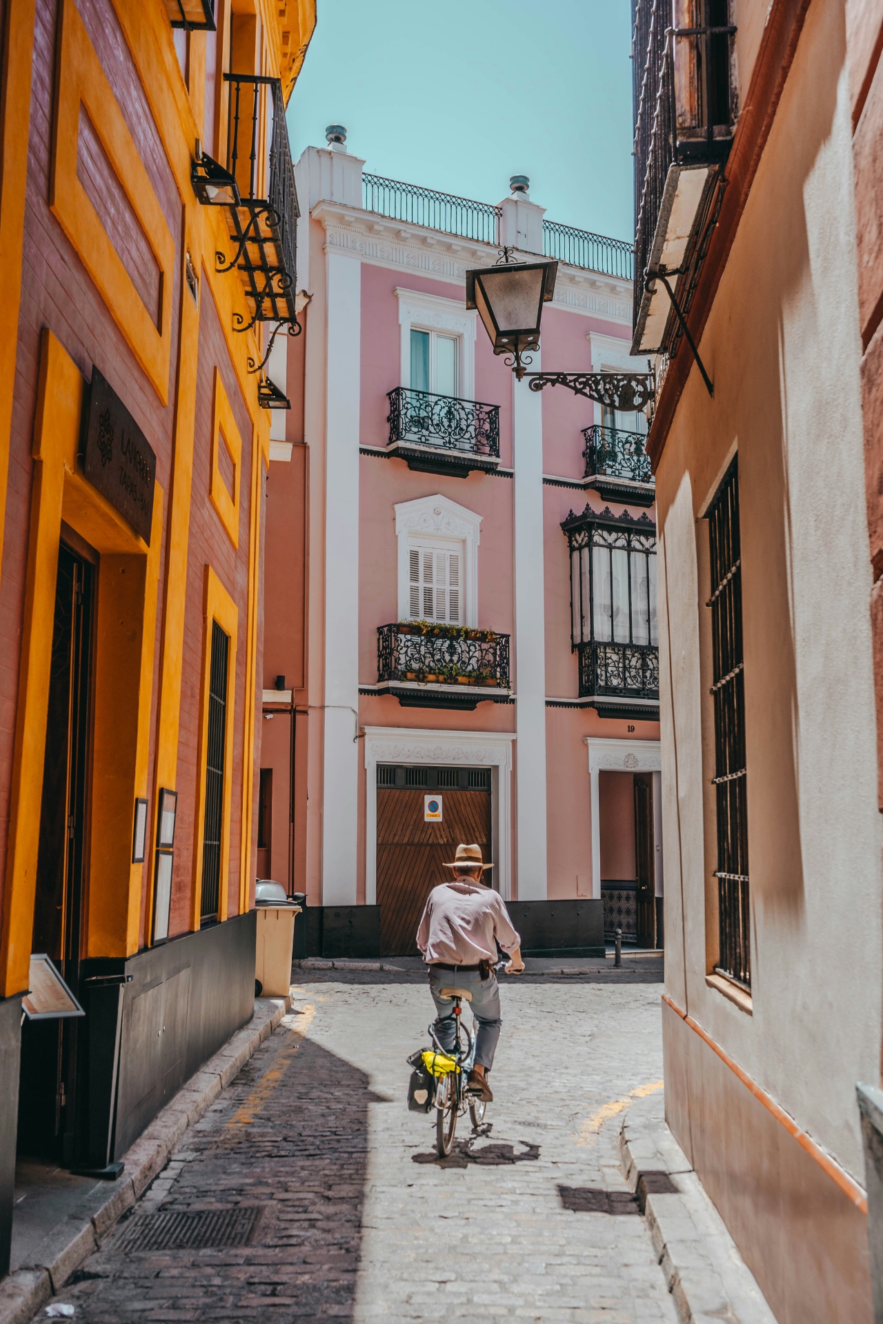 A man in a straw hat cycling down a sunlit narrow Seville street lined with ochre and pink buildings, wrought-iron balconies and a hanging lantern