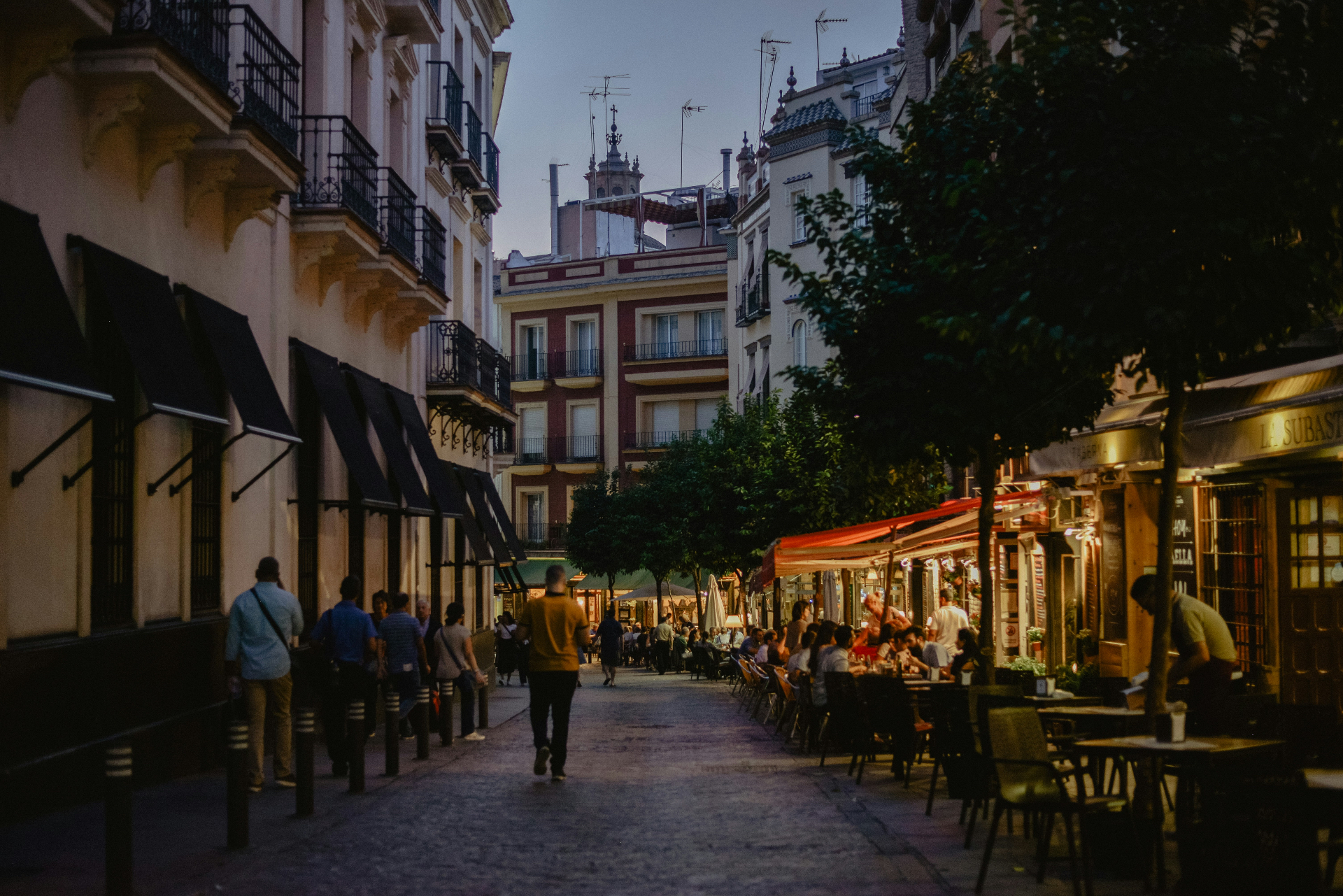 A lively Seville street at dusk with outdoor café tables, lanterns and people gathered along a cobbled lane